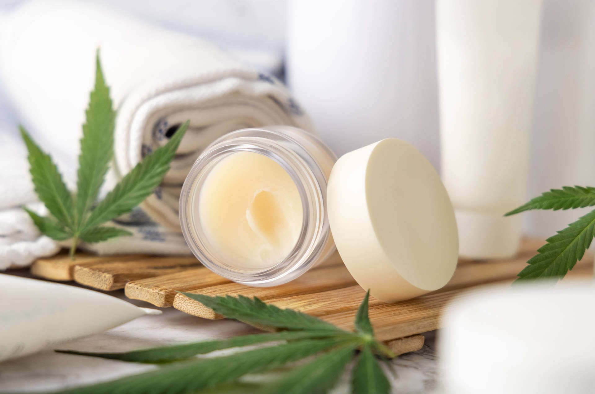 Jar with CBD lip balm and green cannabis leaves near bottles and towel in bathroom close up on a wooden tray. Organic skincare product. Alternative cosmetics